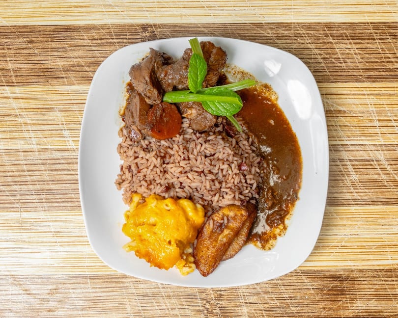 A plate of brown stew chicken, rice and peas, macaroni and cheese, and fried plantain.