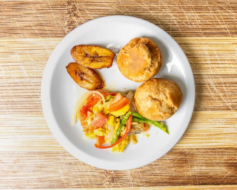 A white plate with fried plantains, two johnny cakes, and ackee and saltfish.