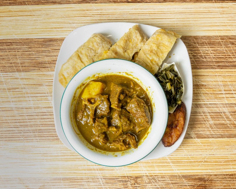 Curried meat and potatoes in a bowl, with roti, collard greens, and fried plantain on a plate.