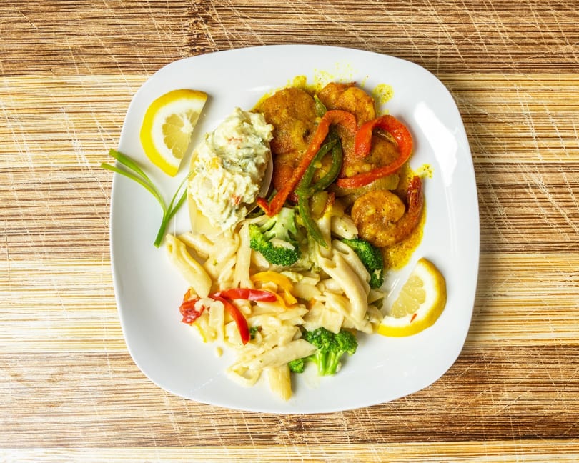 A white plate with shrimp, pasta, broccoli, bell peppers, and potato salad, garnished with lemon.